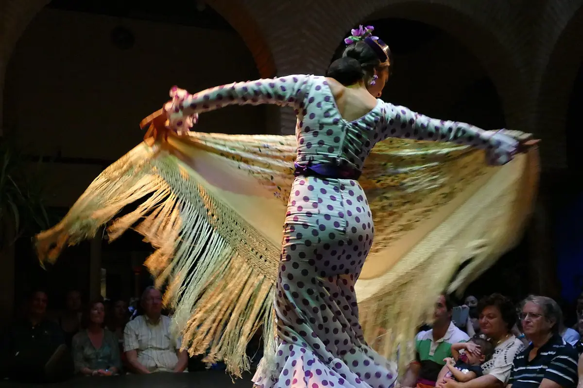 A flamenco dancer in a polka-dot dress swirls a long fringed shawl on a dark stage while an audience watches from close by