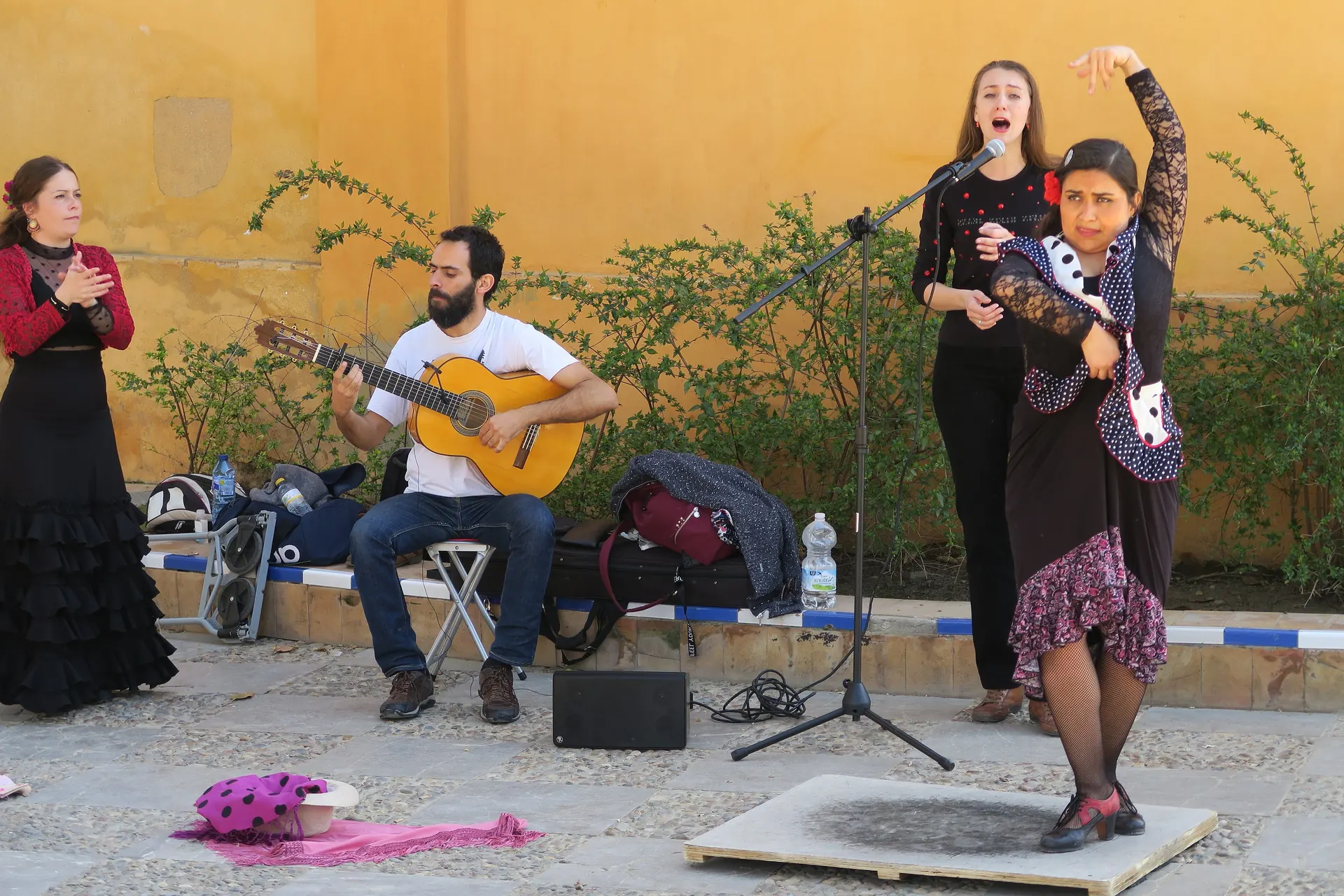 A flamenco dancer in a polka-dot jacket raises one arm at the right, with a singer, a guitarist and a clapping dancer beside her against an ochre wall in Seville