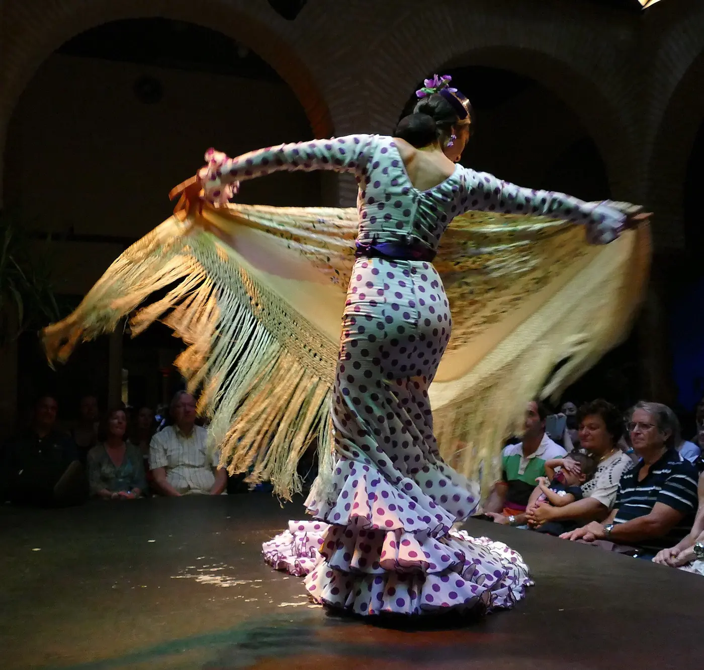 A flamenco dancer in a polka-dot dress swirls a long fringed shawl on a stage while an audience watches from close by